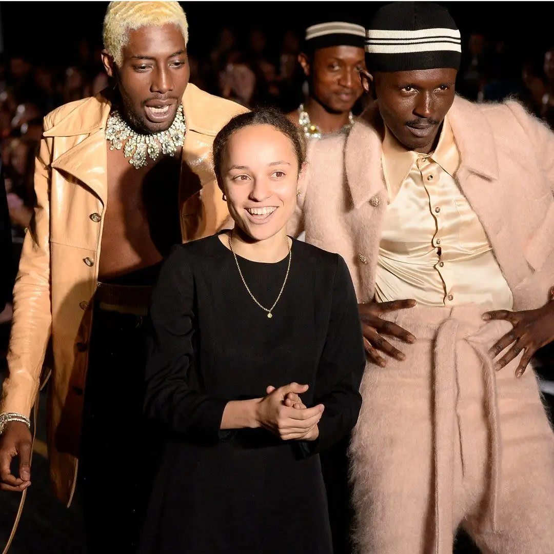 Front-row fashion show moment featuring a smiling Grace Wales Bonner in a simple black dress standing between two models dressed in coordinated neutral-toned looks. One model wears a glossy tan jacket with a statement necklace, while the other is styled in a soft beige ensemble with a fur-textured coat and matching hat. The scene is warmly lit, with a blurred audience in the background, capturing an intimate, celebratory runway atmosphere
