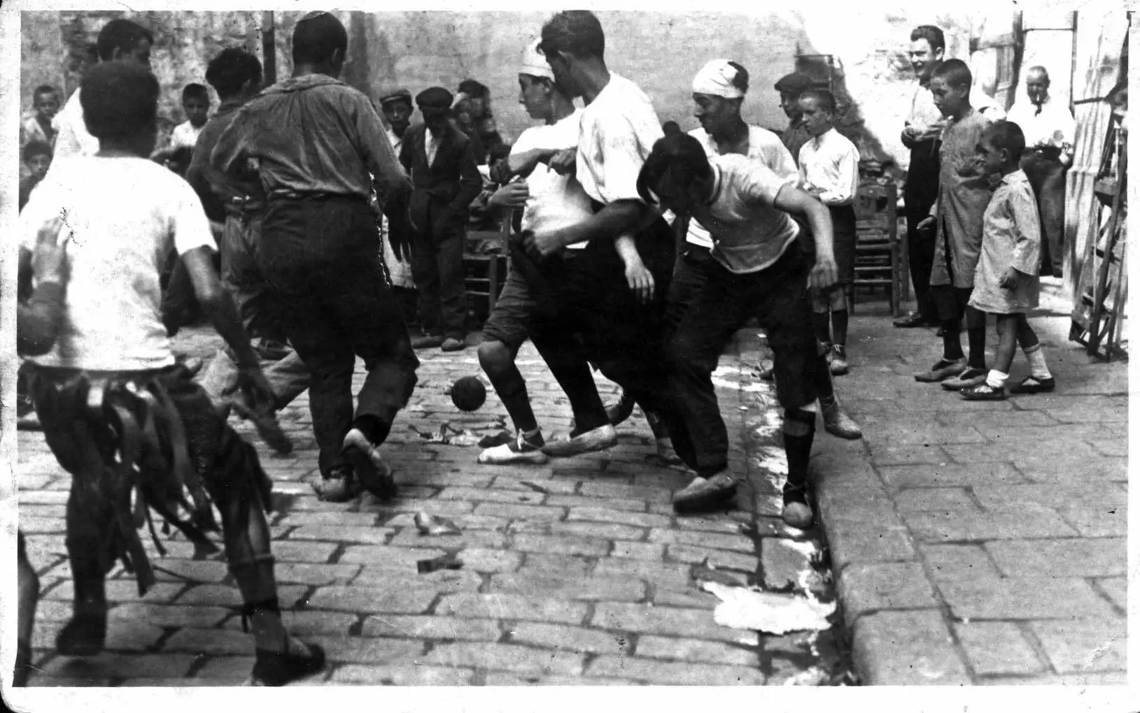 Black-and-white photo of boys playing street football on a cobblestone road, surrounded by onlookers