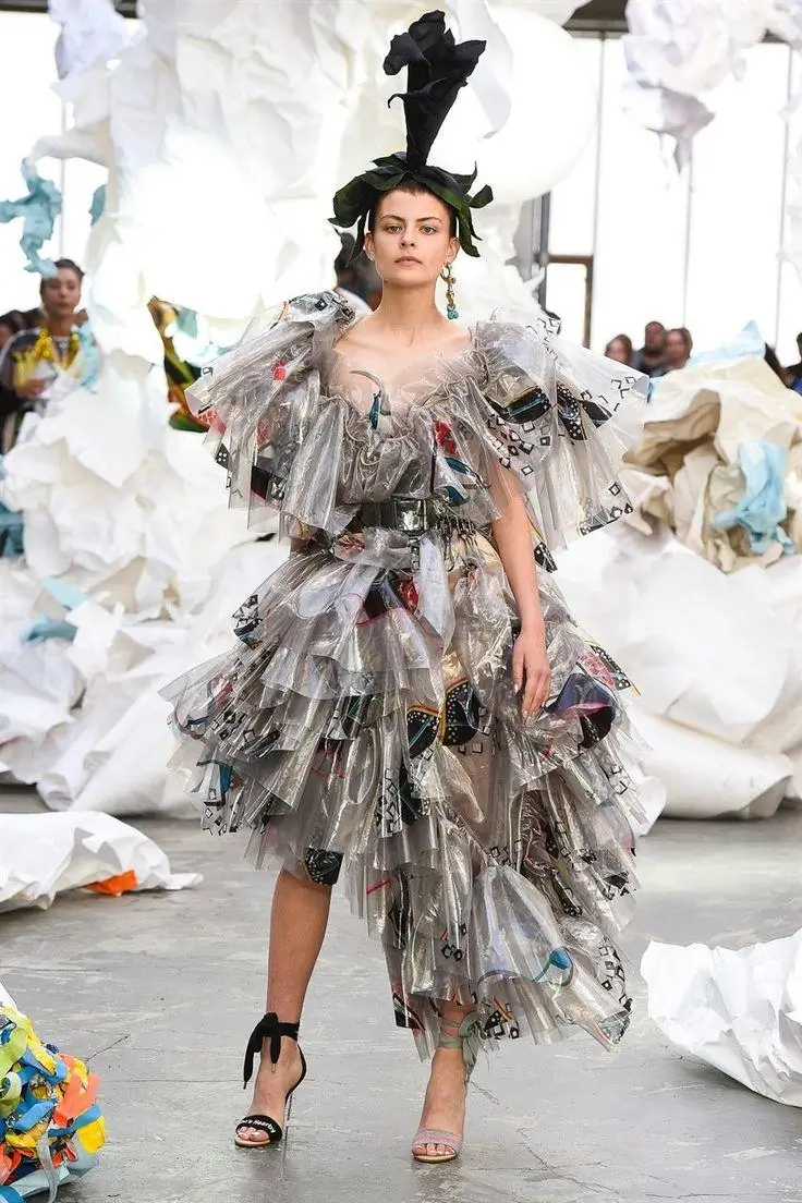 Runway model wearing an avant-garde layered dress made of translucent plastic-like materials with printed fragments, styled with a sculptural headpiece and high heels, set against an installation of oversized crumpled white forms