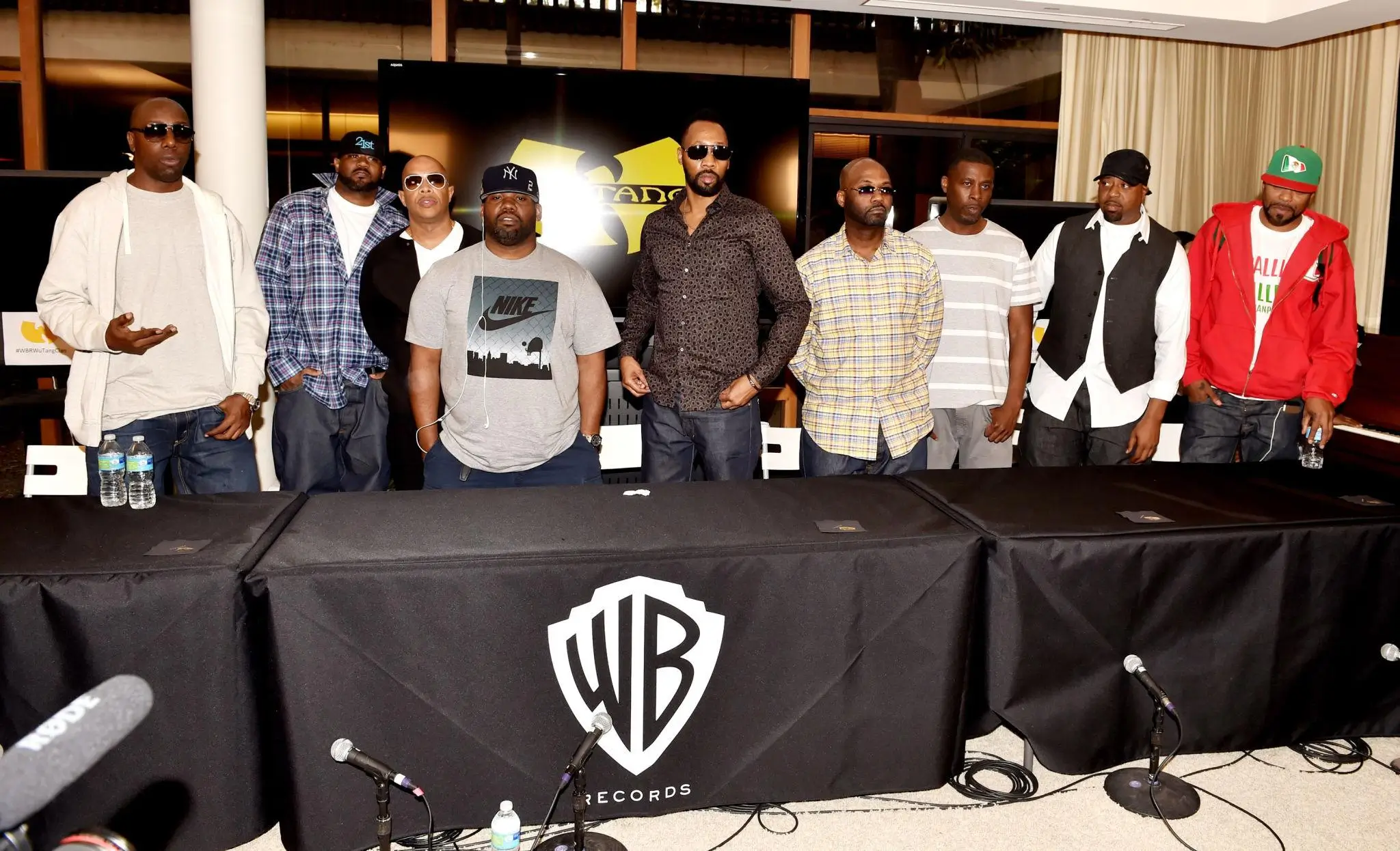 Members of the Wu-Tang Clan stand together behind a table at a press event, dressed in casual streetwear and sunglasses, with a Wu-Tang logo displayed on a screen behind them and a Warner Bros. Records table setup with microphones in front