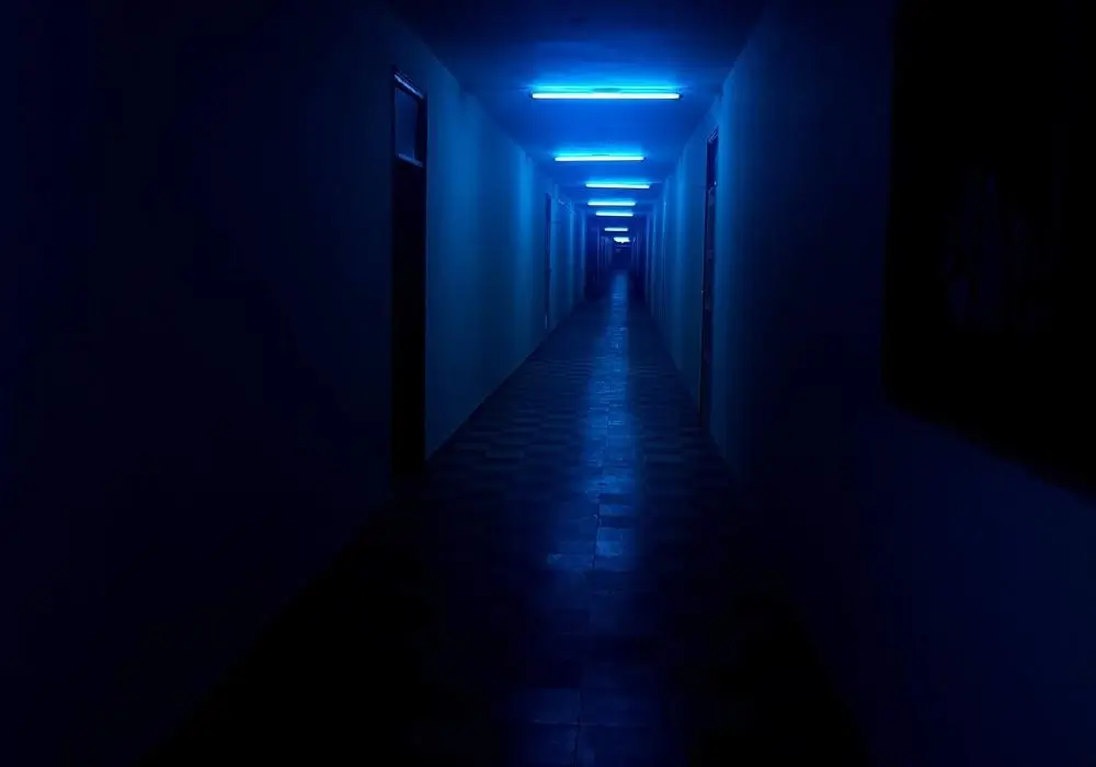 A long, narrow corridor bathed in cold blue fluorescent light stretches into darkness, with evenly spaced ceiling fixtures casting a clinical glow over tiled floors and shadowed doorways, creating an atmosphere of isolation and psychological tension