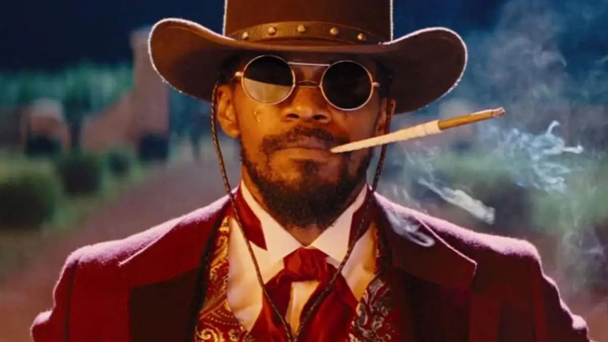 A sharply dressed man styled like a Western gunslinger stands outdoors in warm, cinematic light, wearing a wide-brimmed cowboy hat, round tinted sunglasses, and a deep red tailored suit with a patterned vest and tie. A cigarette rests at the corner of his mouth, trailing smoke into the air, while his composed expression and upright posture project calm authority against a softly blurred frontier-town backdrop