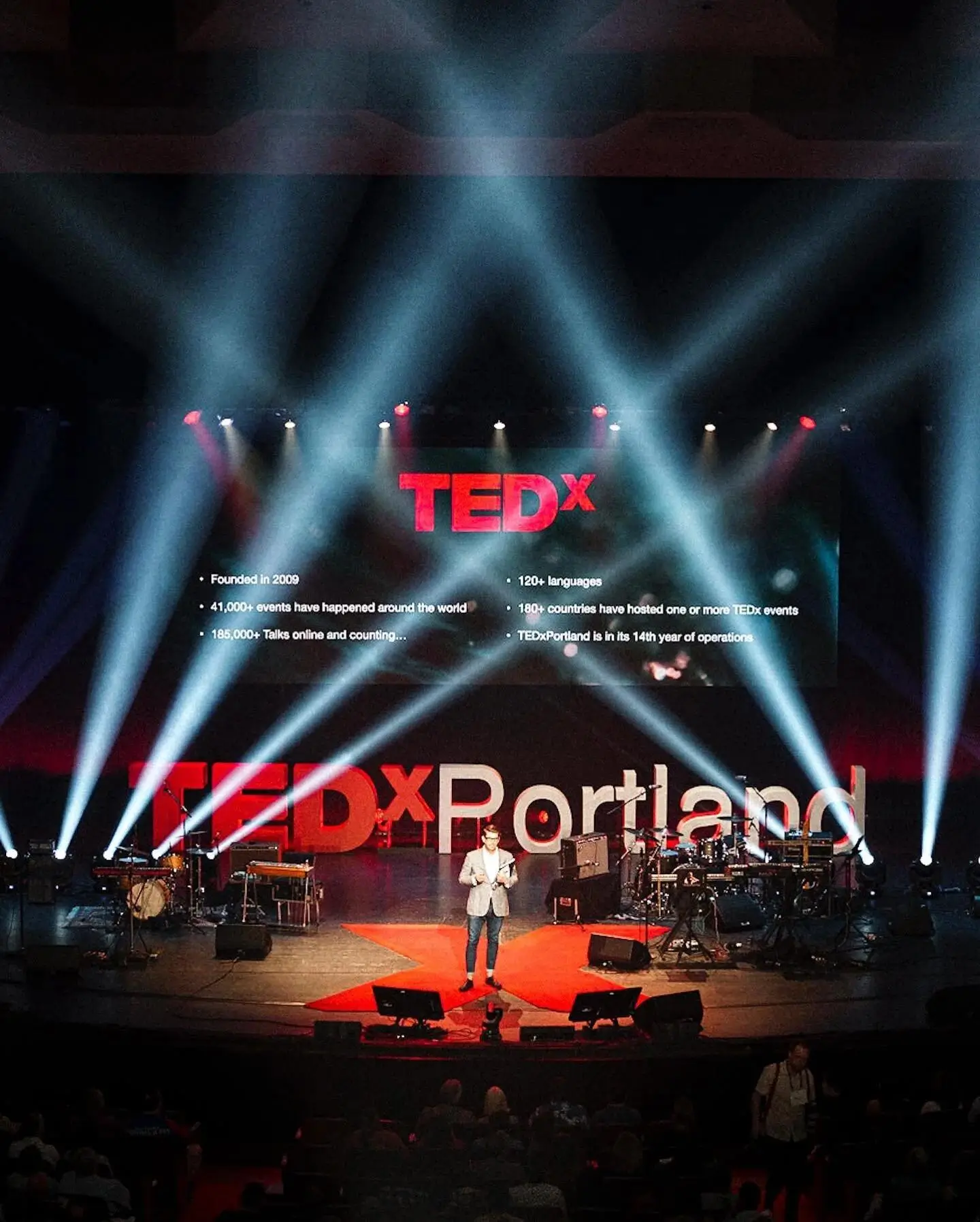 A wide-angle view of a live TEDx Portland event shows a speaker standing center stage on the iconic red circular carpet, delivering a talk beneath dramatic beams of white and blue stage lighting. Behind them, a large screen displays the TEDx logo alongside key statistics about the platform—its founding in 2009, global reach across 180+ countries, and tens of thousands of events and talks. The stage is flanked by musical instruments and equipment, suggesting a multidisciplinary program that blends talks with live performance. In the foreground, an audience sits in a dimly lit auditorium, focused on the presentation, while the bold “TEDxPortland” lettering stretches across the back of the stage, anchoring the event’s identity