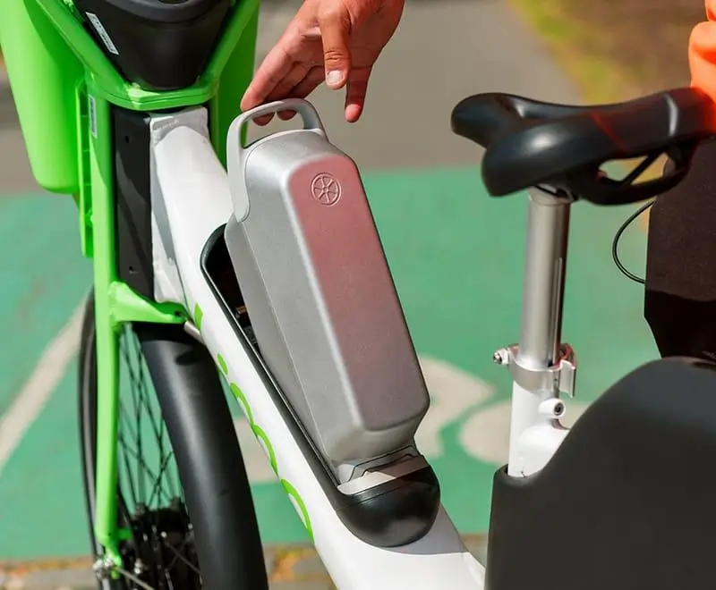 Hand inserting a removable battery into the rear-mounted housing of a Lime electric bike, highlighting the updated battery placement behind the seat post for improved balance and accessibility