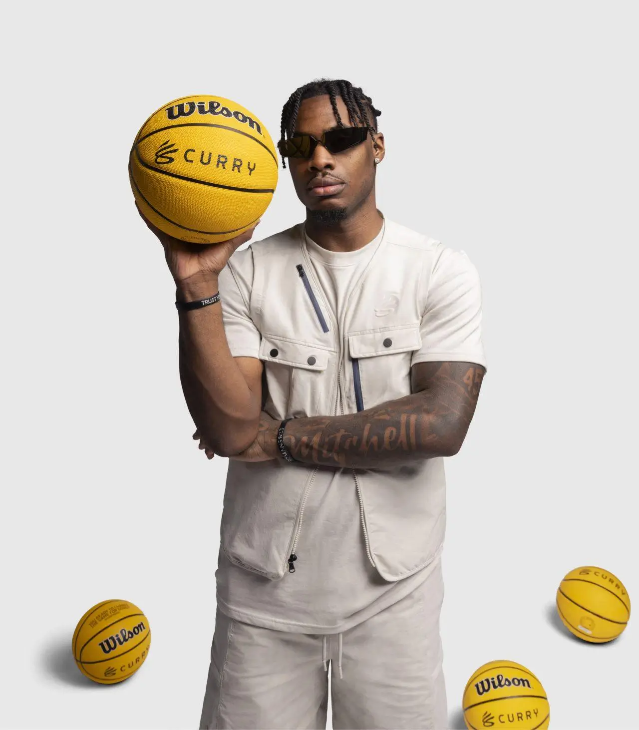 Davion Mitchell wearing neutral-toned vest and sunglasses holding a Wilson basketball with Curry branding against a studio background