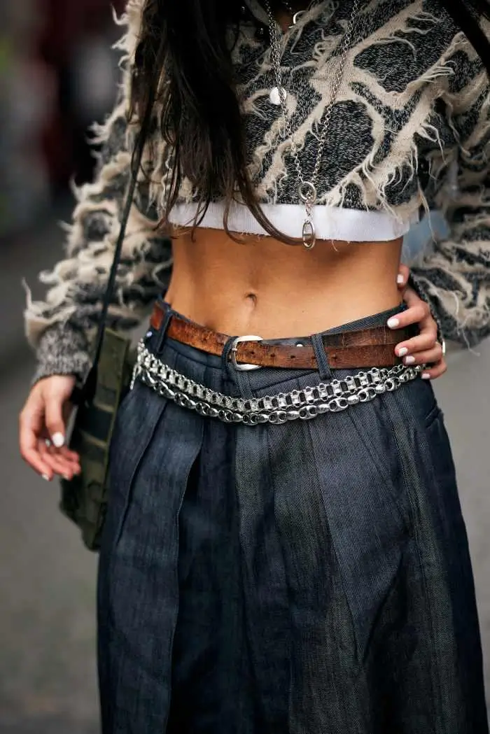 Close-up, waist-level shot focusing on a layered, textural outfit. The subject wears a cropped knit top with a distressed, frayed pattern in neutral tones, exposing the midriff and emphasizing a raw, deconstructed aesthetic. Around the neck, multiple silver chain necklaces of varying lengths add a metallic contrast, with one longer pendant resting near the waistline. The lower half features high-waisted, dark pleated trousers with a soft drape, structured yet relaxed. At the waist, styling becomes the focal point: a worn brown leather belt sits above a layered silver chain belt that wraps loosely across the hips, introducing movement and an industrial edge. One hand rests casually near the pocket, while the other holds a bag with a subtle camo pattern, reinforcing the mix of utilitarian and street-inspired elements. Overall, the look blends grunge-inspired textures with tailored structure, anchored by accessories that create depth and visual tension