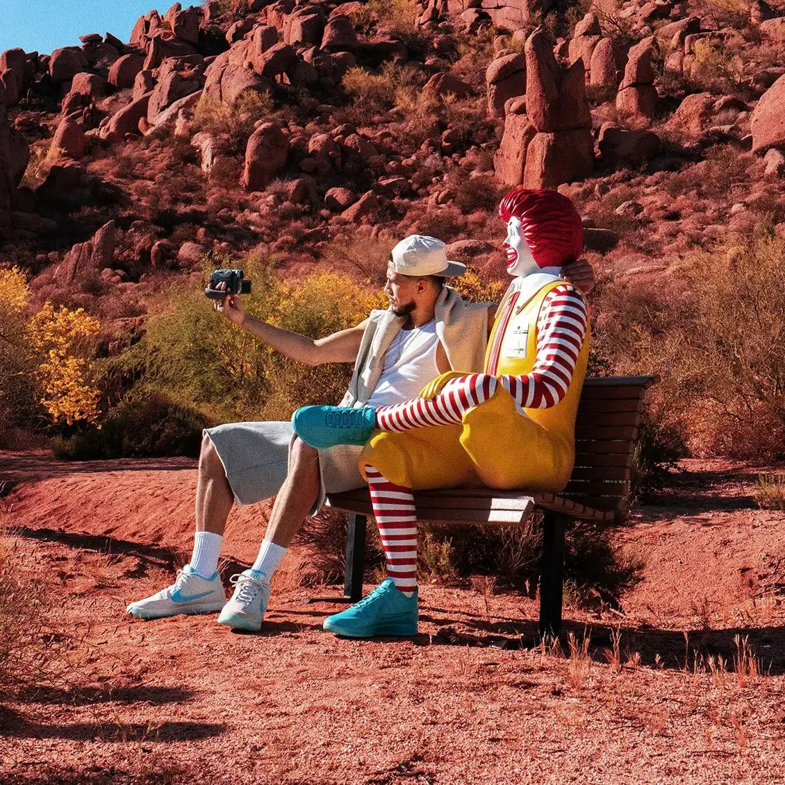 Desert campaign scene featuring a person seated beside the Ronald McDonald mascot in rocky Arizona terrain while showcasing the McDonald’s x Nike Book 2 “Sedona” collab