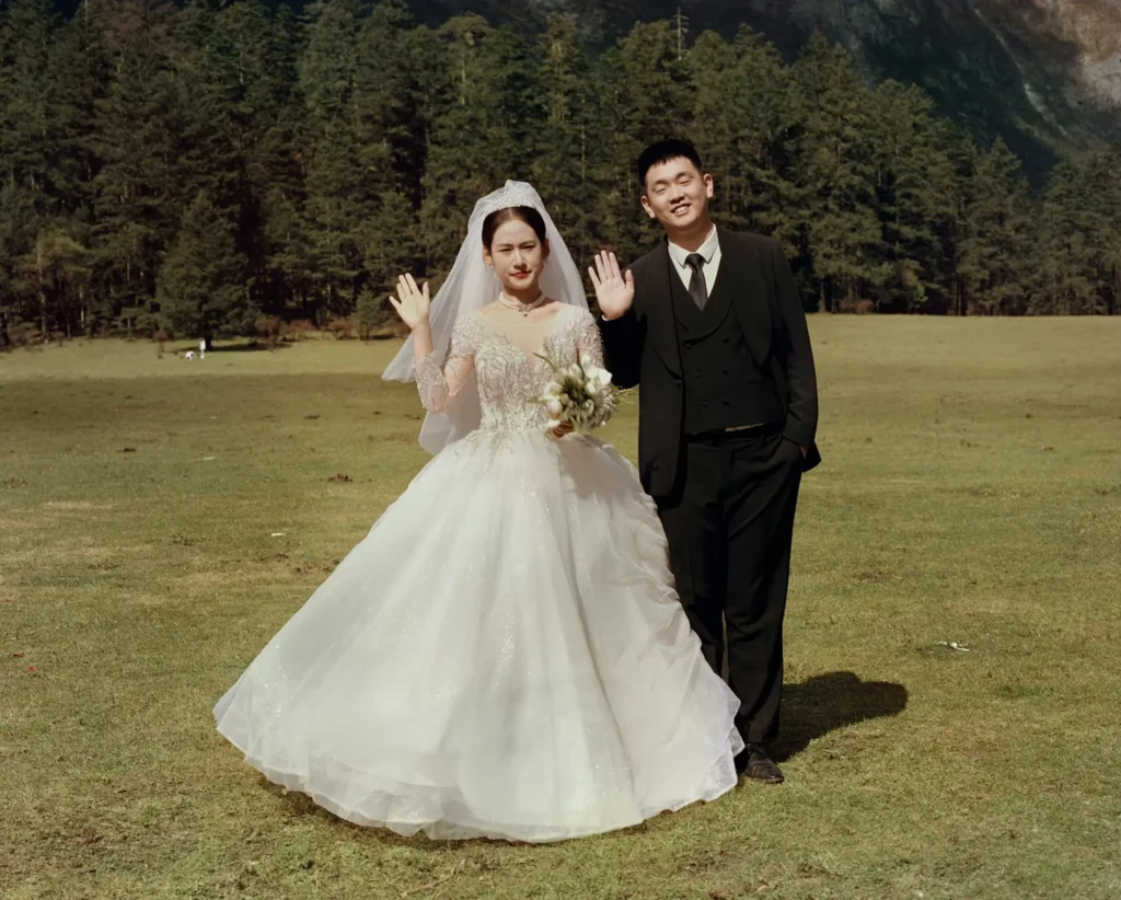 A bride in an ornate white wedding gown and a groom in a black suit stand in an open alpine meadow waving toward the camera, framed by dense evergreen forests and dramatic mountain scenery