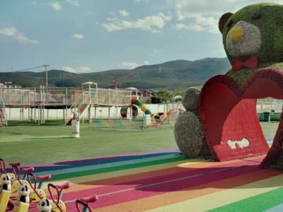A brightly tinctured outdoor playground features rainbow flooring, children’s tricycles, and a giant bear-shaped play structure holding a red heart, set against rolling mountain landscapes under a clear sky