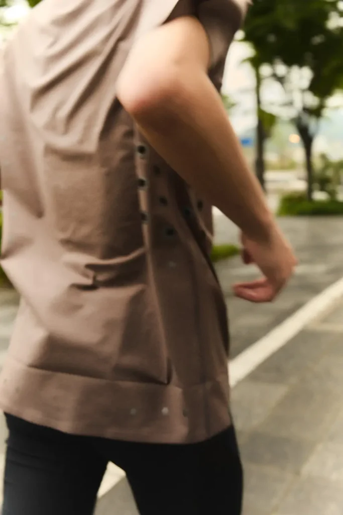 Rear-side view of a runner wearing a muted brown technical performance T-shirt with subtle perforated ventilation details along the side panel, captured mid-stride on a quiet urban running path lined with trees in soft evening light