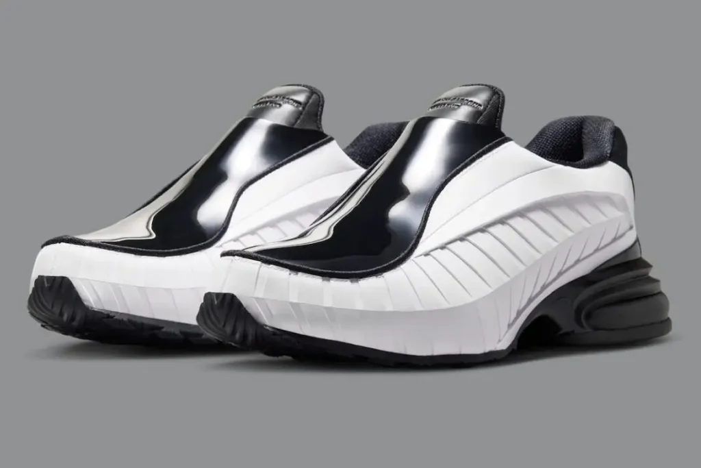A pair of COMME des GARÇONS HOMME PLUS x Nike Air Max Dolce shoe shown in a black-and-white colorway against a neutral gray background. The avant-garde slip-on silhouette features glossy patent leather uppers, sculpted ribbed sidewalls, and a futuristic chunky heel with visible Air-inspired cushioning. The laceless construction and exaggerated proportions blend luxury formalwear aesthetics with early-2000s Nike performance design, creating a sleek sneaker-loafer hybrid appearance