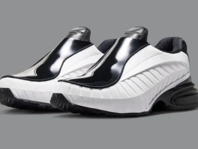 A pair of COMME des GARÇONS HOMME PLUS x Nike Air Max Dolce shoe shown in a black-and-white colorway against a neutral gray background. The avant-garde slip-on silhouette features glossy patent leather uppers, sculpted ribbed sidewalls, and a futuristic chunky heel with visible Air-inspired cushioning. The laceless construction and exaggerated proportions blend luxury formalwear aesthetics with early-2000s Nike performance design, creating a sleek sneaker-loafer hybrid appearance