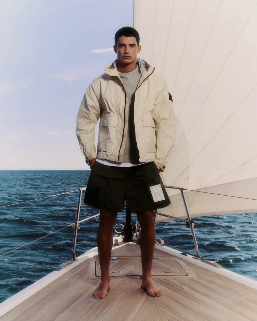 A model stands barefoot at the bow of a sailboat, framed by open ocean and a towering white sail under soft daylight. He wears a light cream technical hooded jacket with utilitarian flap pockets and a signature sleeve patch, layered over a muted grey top and paired with black wide-leg shorts featuring a contrasting rectangular patch detail near the hem. The composition blends luxury sportswear with maritime minimalism, capturing a calm yet structured summer atmosphere through clean tailoring, wind-resistant textures, and muted coastal tones
