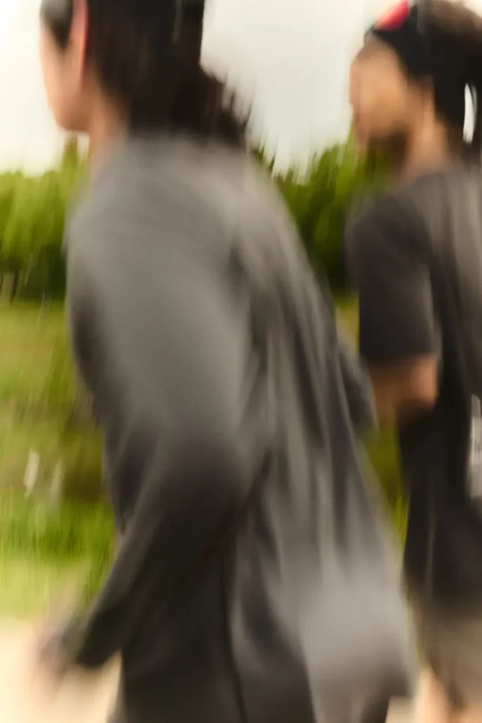 Motion-blurred photograph of two runners moving side by side along an outdoor trail, wearing dark technical performance layers and athletic apparel against a soft green landscape backdrop in warm natural light