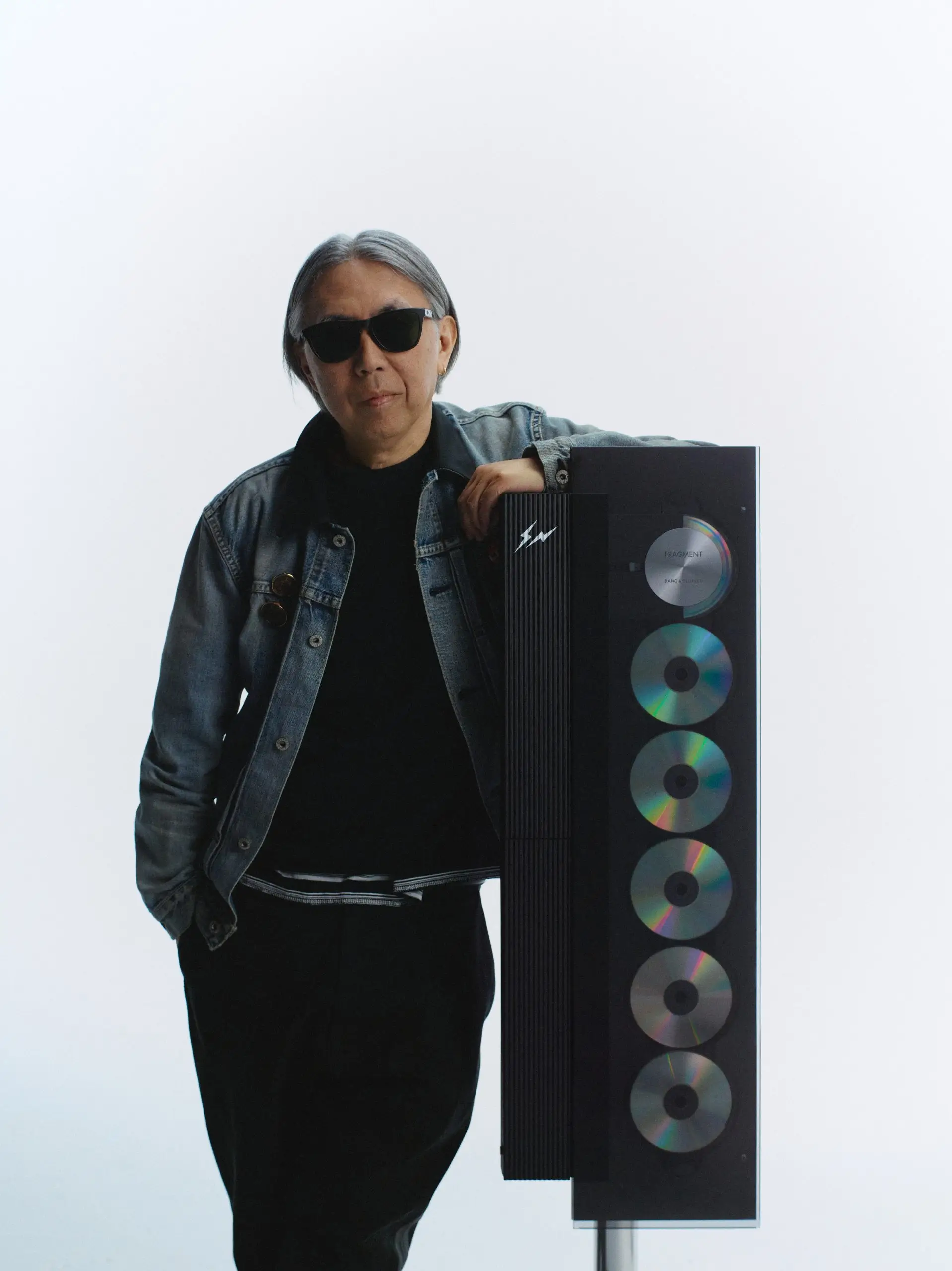 A minimalist campaign portrait featuring a person standing beside a vertically displayed luxury audio system in a clean white studio setting. The subject wears dark sunglasses, a faded indigo denim jacket layered over black essentials, and stands with one hand in a pocket while resting the other casually on the audio unit. The glass-front system reveals a vertical sequence of reflective compact discs with subtle iridescent color shifts, contrasted by a ribbed matte-black side panel carrying a discreet lightning-bolt insignia. The composition balances understated streetwear styling with precision industrial design, reinforcing themes of quiet luxury, monochrome refinement, and collectible audio culture