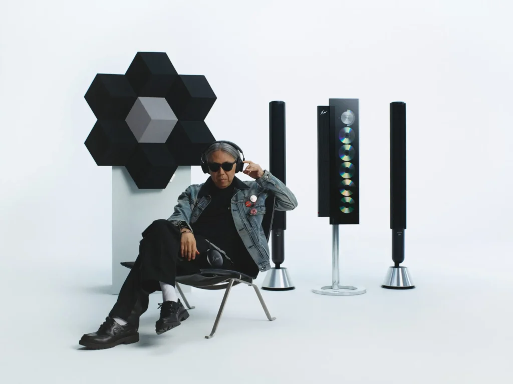 A minimal campaign portrait featuring a seated person wearing black Bang & Olufsen over-ear headphones and dark sunglasses, positioned within a gallery-like white studio environment. Dressed in a faded denim jacket layered over dark clothing, the subject sits casually in a low lounge chair while adjusting the headphones. Surrounding them is the full Bang & Olufsen x Fragment Design collection: a geometric modular speaker installation with an optical cube effect mounted on a pedestal, a vertically displayed glass-front audio system revealing stacked compact discs, and a pair of slim floor-standing speakers in matching monochrome finishes. The composition blends luxury audio, contemporary furniture, and understated streetwear sensibility into a study of quiet design and immersive listening culture