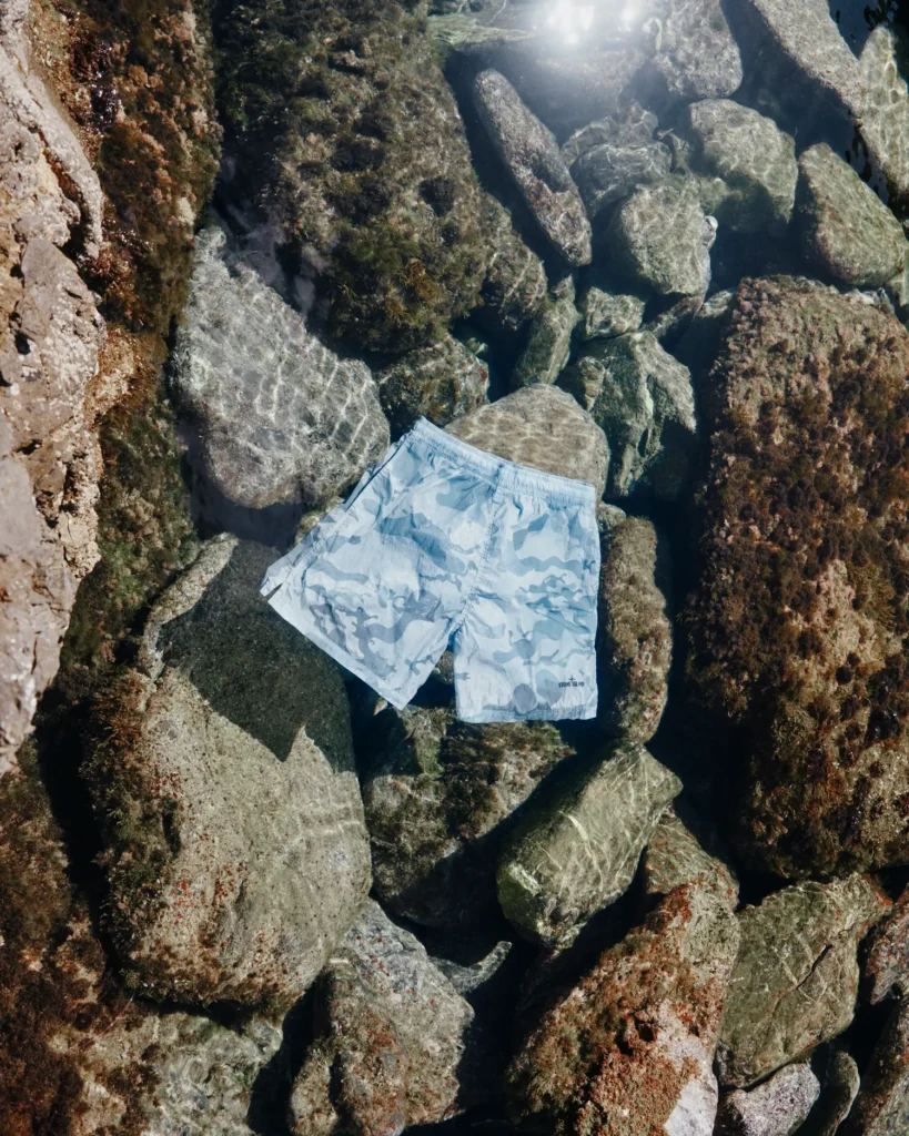 A pair of pale blue camouflage swim shorts rests beneath crystal-clear seawater among rugged coastal rocks and sea-worn textures. Sunlight filters across the surface, creating shimmering reflections and rippling distortions that amplify the garment’s washed aquatic palette. The composition blends technical resortwear with natural Mediterranean scenery, emphasizing Stone Island’s balance of utility, maritime identity, and understated summer luxury through camouflage graphics, lightweight fabrication, and environmental immersion