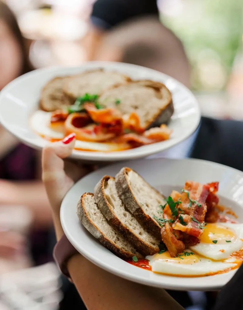 Close-up of two hands holding shallow white plates of brunch food, featuring sunny-side eggs in a rich tomato sauce topped with crispy bacon and fresh herbs, served alongside thick slices of rustic artisan bread, with a softly blurred café setting in the background