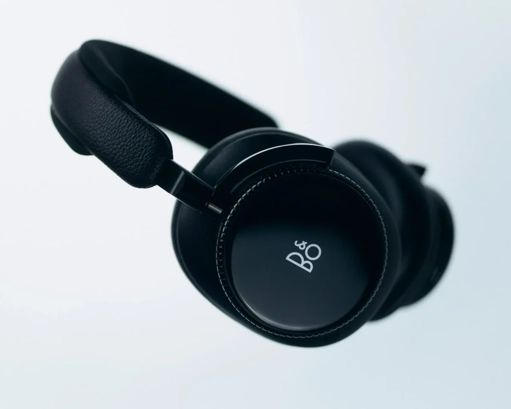 A pair of premium over-ear headphones presented in an ultra-clean studio composition, finished entirely in deep black tones with subtle satin and matte contrasts. The design features softly cushioned ear cups, a textured leather headband, and a precision-machined circular outer shell marked only by a discreet Bang & Olufsen logo. Fine ridged detailing surrounds the ear cup edge, emphasizing tactile control and refined engineering. Suspended against a pale minimal backdrop, the headphones convey a balance of luxury audio craftsmanship, contemporary restraint, and understated monochrome elegance