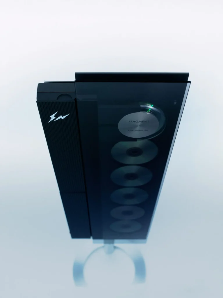 A vertically mounted luxury audio system shown in an ultra-minimal studio setting, finished in deep monochrome black with a glossy glass front panel. Behind the tinted surface, a vertical stack of compact discs is visible, while one disc is actively suspended and illuminated with subtle green light accents during playback. The left side features ribbed detailing and a discreet white Fragment Design lightning-bolt insignia, contrasting against the otherwise stealth-like finish. Elevated on a slim stand, the object blends vintage hi-fi mechanics with contemporary sculptural design, reading as both precision audio equipment and gallery-worthy industrial art