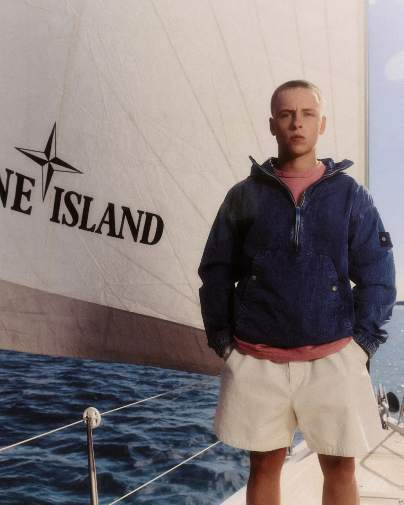 A model stands aboard a sailing yacht against a massive white sail marked with the Stone Island compass insignia, surrounded by deep blue open water and warm late-day light. He wears a navy technical pullover jacket with a half-zip construction and signature sleeve badge, layered over a faded rose crewneck and paired with relaxed off-white shorts. The image balances nautical elegance with functional sportswear, combining washed textures, muted summer tones, and maritime minimalism to evoke a refined coastal lifestyle rooted in utility and contemporary luxury