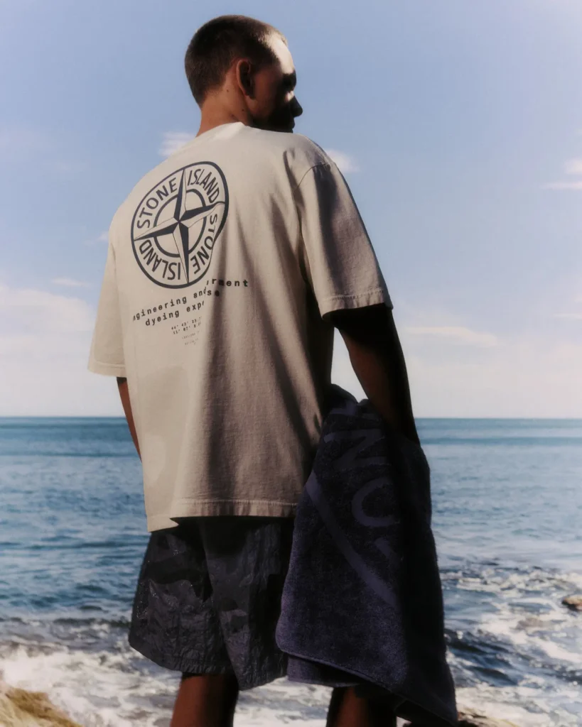 A model stands along a rocky shoreline overlooking calm open water beneath a clear sky, dressed in relaxed coastal essentials from Stone Island. He wears an oversized light beige graphic T-shirt featuring an enlarged compass logo and technical-inspired typography across the back, paired with dark lightweight shorts and carrying a navy towel draped casually at his side. Soft sunlight and ocean reflections enhance the washed palette and understated maritime atmosphere, blending casual resortwear with Stone Island’s signature fusion of functional design, garment experimentation, and contemporary
