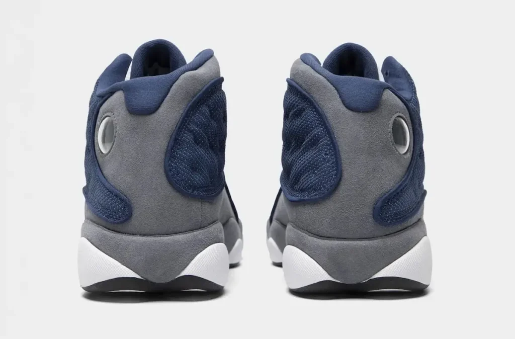 Rear view of the Air Jordan 13 featuring grey suede heel panels, navy quilted mesh overlays, padded navy collars, and signature circular holographic emblems near the ankle, set against a min light grey studio backdrop