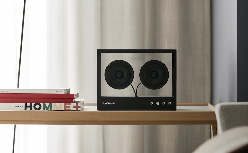 Transparent Sound Small Speaker in Black positioned on a light wooden table beside stacked design magazines within a soft neutral-toned Scandinavian interior, featuring exposed dual drivers and minimalist industrial styling