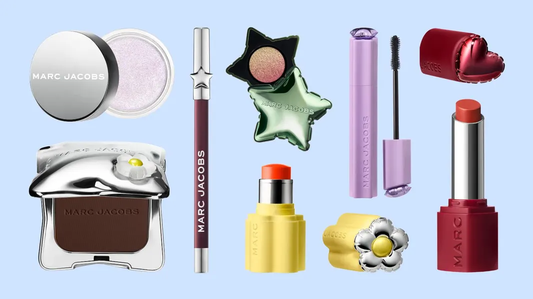 Marc Jacobs Beauty’s 2026 collection displayed against a soft blue background, featuring colorful makeup essentials including mascara, eyeliner, lipstick, bronzer, highlighter, blush stick, and eyeshadow. The products showcase play heart, flower, star, and chrome-inspired packaging that blends whimsical design with modern haute