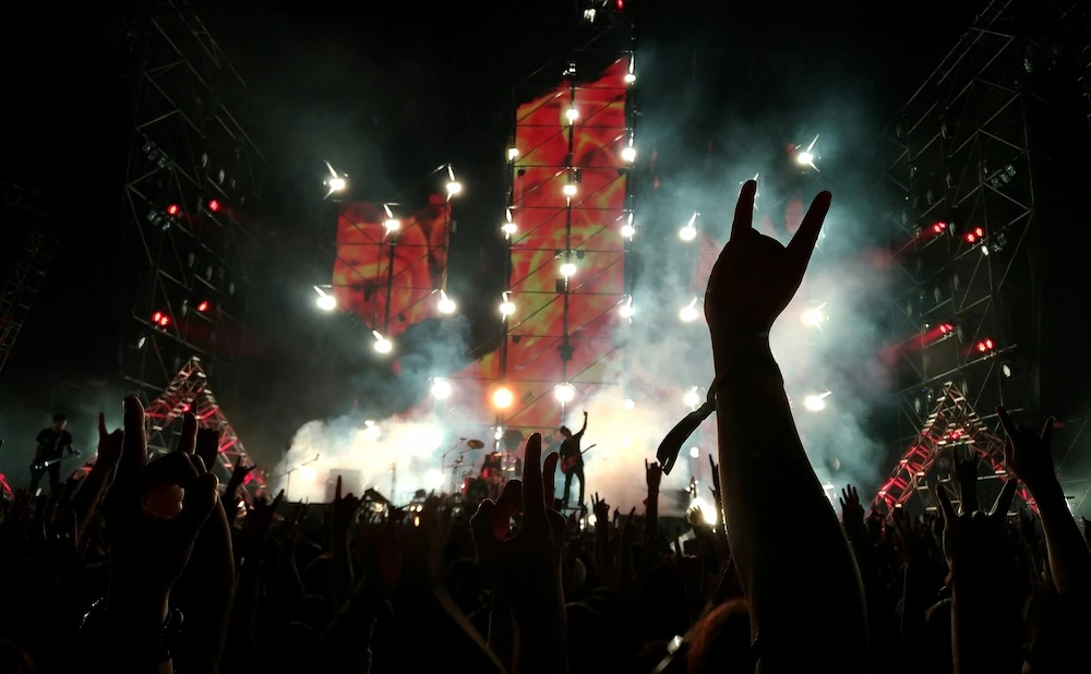 Crowd at a nighttime rock concert with raised hands forming horn gestures, silhouetted against bright stage lights, smoke effects, and towering illuminated screens. A performer stands center stage amid dramatic red and white lighting, creating an energetic festival atmosphere