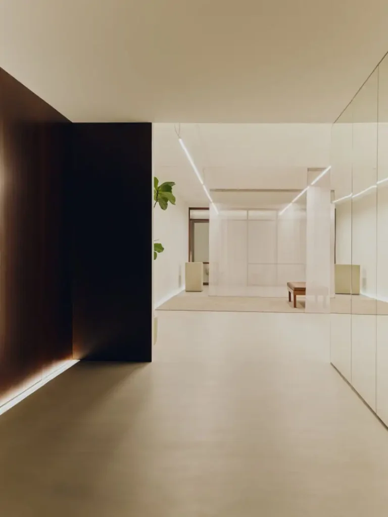Minimalist Mediterranean-inspired interior space with warm ambient lighting, featuring dark wood paneling, reflective glass walls, soft translucent curtains, and a clean open-plan layout. Linear ceiling lights stretch through the serene architectural environment, while neutral tones, polished flooring, and sparse furnishings create a quiet, gallery-like atmosphere rooted in contemporary haute design