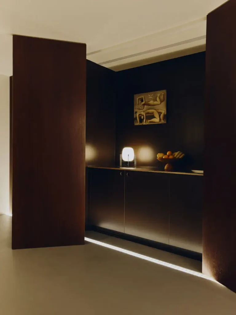 Warmly lit contemporary interior featuring rich dark wood paneling, minimalist cabinetry, and soft architectural lighting that glows along the floor and ceiling edges. A sculptural table lamp, ceramic fruit bowl, and textured wall artwork introduce subtle Mediterranean character, while the restrained palette and clean spatial composition create an intimate, gallery-like atmosphere rooted in modern haute and quiet design precision