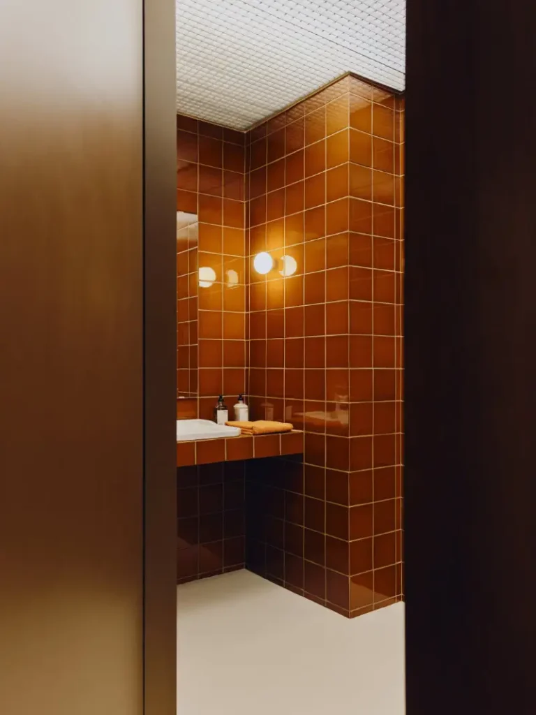 Intimate modern bathroom interior framed by dark wood surfaces and glossy caramel-brown tiled walls, illuminated by soft spherical wall lights that cast a warm ambient glow. Minimalist fixtures, folded towels, and clean geometric lines create a refined Mediterranean-inspired atmosphere, blending tactile materiality with understated contemporary