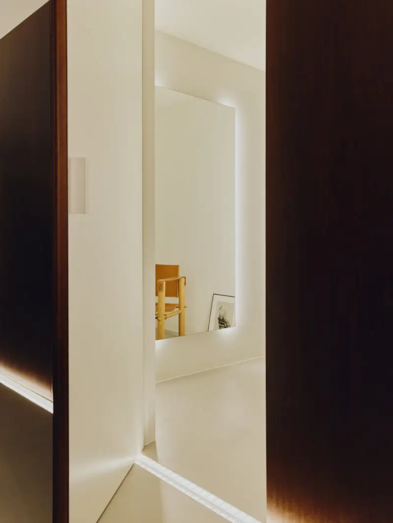 Minimal contemporary interior with warm ambient lighting, featuring dark wood paneling, soft cream walls, and a glowing backlit mirror that enhances the serene architectural composition. A simple wooden chair and framed artwork subtly emerge within the reflective space, while concealed floor lighting and restrained material textures create a calm Mediterranean-inspired atmosphere rooted in quiet and spatial precision