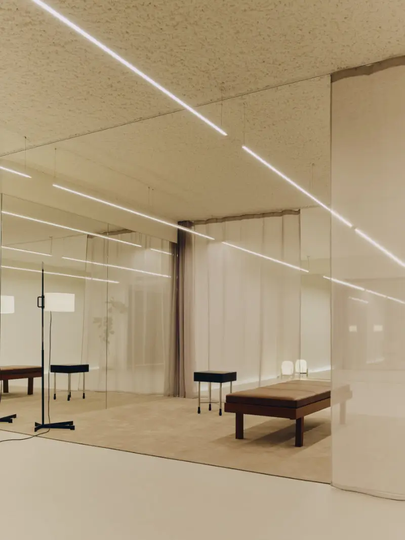 Minimalist gallery-like interior featuring mirrored walls, sheer floor-to-ceiling curtains, and long linear ceiling lights that reflect across the space with soft symmetry. Sparse furnishings, including a low leather bench and sculptural standing lamp, sit within a warm neutral palette, creating a serene contemporary atmosphere shaped by Mediterranean restraint, architectural precision, and quiet haute