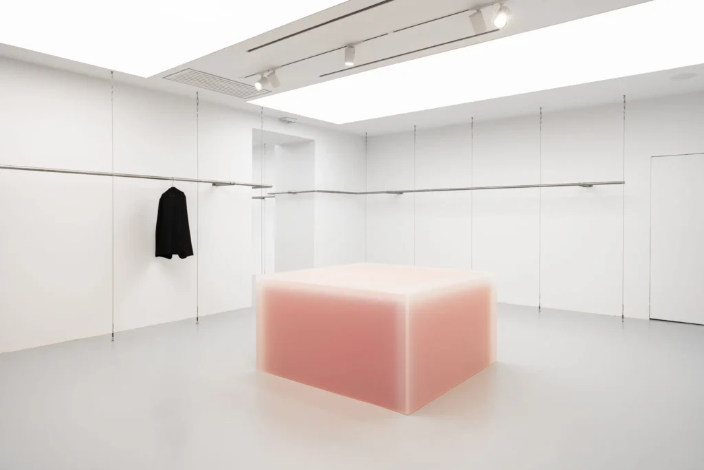 Minimalist concept store interior featuring white walls, suspended metal clothing rails, and a single black garment, centered around a glowing translucent cube display with soft pink illumination and polished concrete flooring