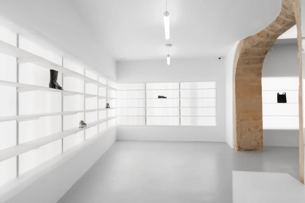 Minimalist concept store interior with continuous white shelving displaying sparse footwear and accessories, soft overhead strip lighting, polished concrete flooring, and a contrasting exposed limestone arch framing the space