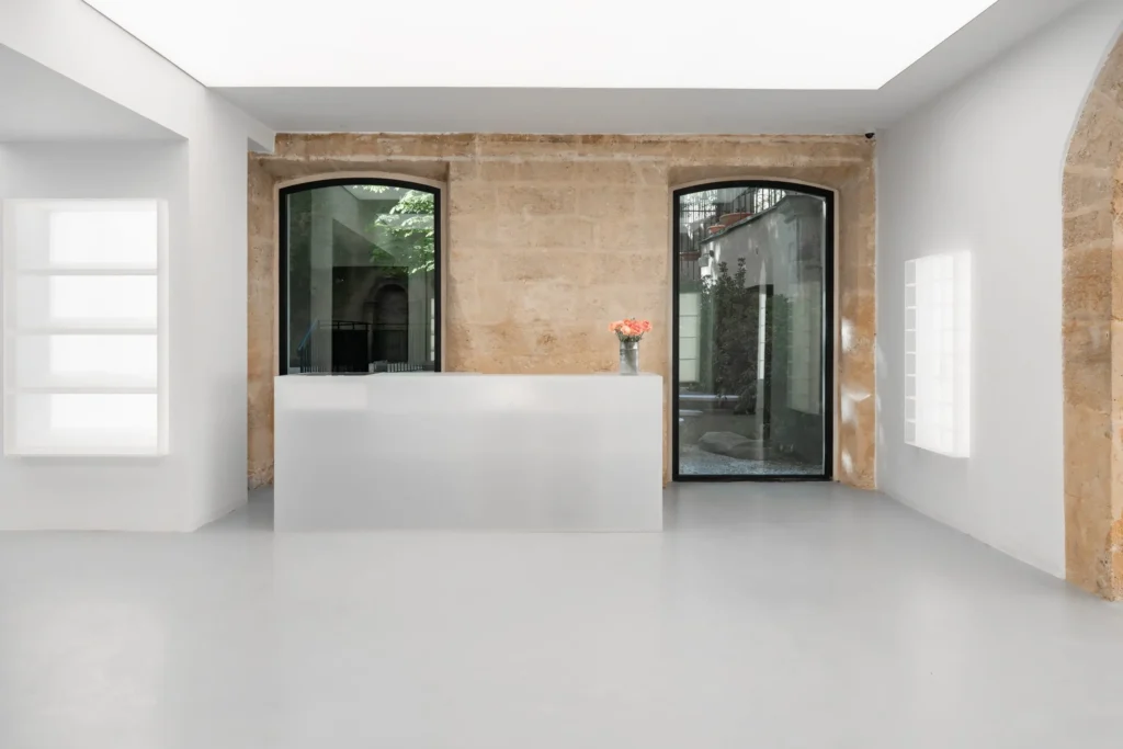 Minimalist concept store interior with a central white reception counter, exposed limestone wall with arched windows, soft natural light, and a small floral arrangement adding contrast to the clean, gallery-like space