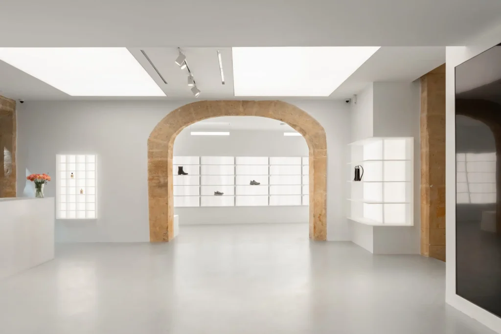 Minimalist concept store interior framed by a large exposed limestone arch, with illuminated white shelving displaying select footwear and accessories, polished concrete flooring, and soft overhead lighting enhancing the gallery-like space