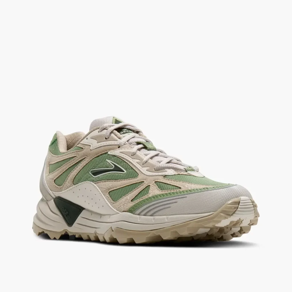 Low-top trail-inspired shoe in muted sage green mesh with cream suede overlays, featuring a rugged lug outsole, sculpted off-white midsole, and subtle dark green branding accents. The layered technical design blends outdoor performance aesthetics with contemporary lifestyle styling against a clean light gray background
