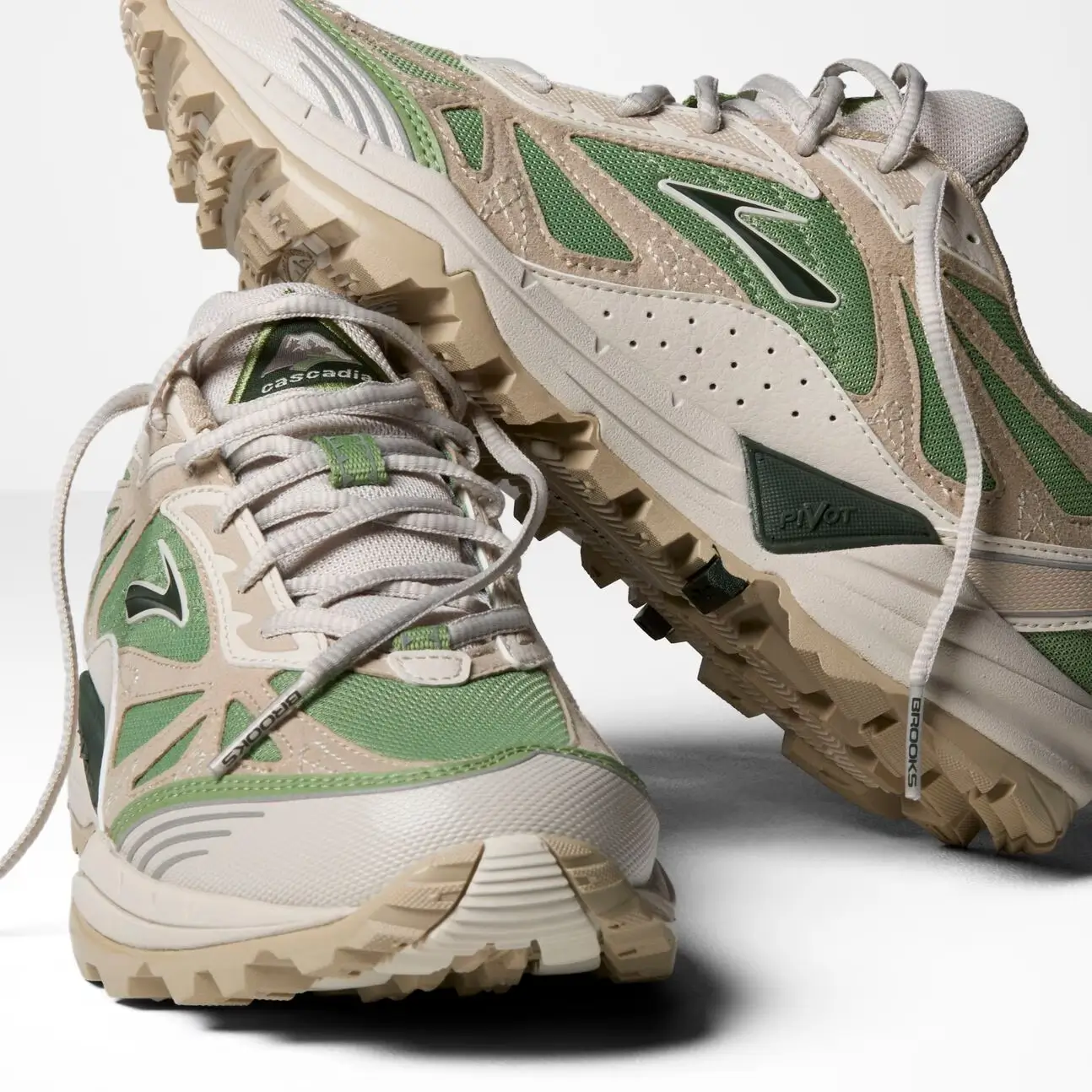 Close-up product shot of Brooks Cascadia trail-inspired sneakers in muted sage green and cream tones, showcasing layered suede overlays, breathable mesh construction, rugged lug soles, and technical outdoor detailing. The chunky off-white midsoles, reflective accents, and trail-ready traction emphasize the shoe’s blend of archival performance heritage and modern lifestyle design against a clean studio backdrop