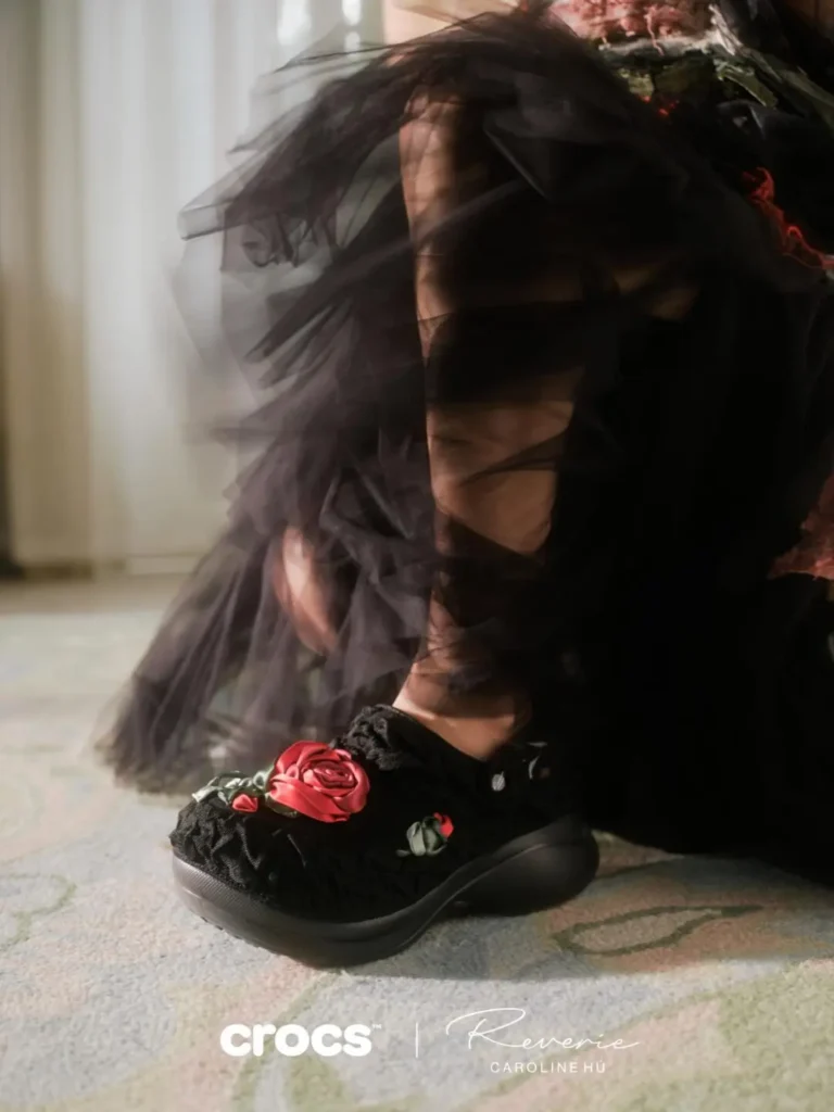 Model wearing black floral Crocs x Caroline Hú Bae Clogs beneath layered sheer black fabric creating a romantic cinematic mood