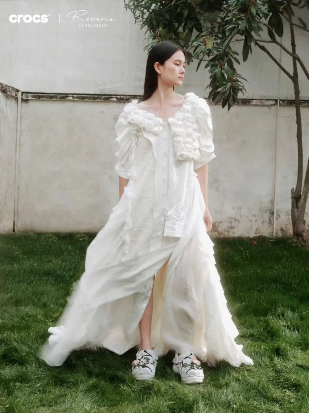Model wearing an ethereal white textured gown outdoors, styled with white Crocs x Reverie by Caroline Hú floral Bae Clogs featuring cracked detailing and soft embroidered blooms against a garden backdrop