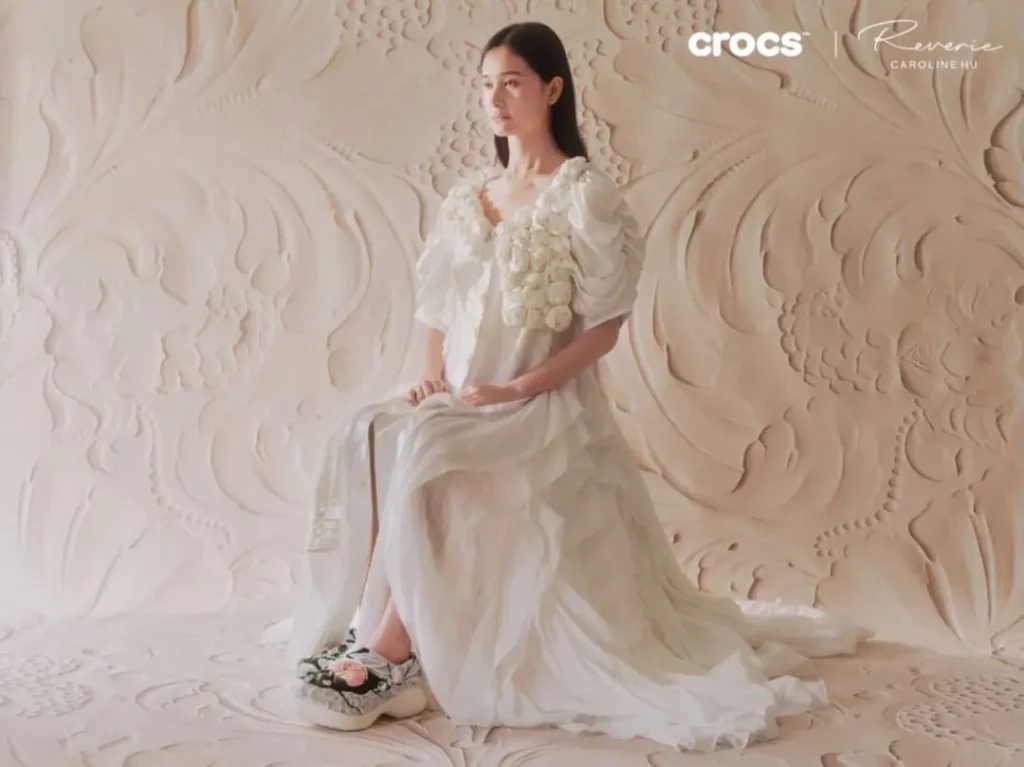 Model seated in sculptural white surroundings wearing embroidered Crocs x Caroline Hú clogs and couture-inspired dress