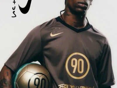 Studio campaign-style portrait of a person wearing a dark Nike football jersey while holding a metallic gold soccer ball marked with the number “90.” The shirt features matching gold graphics, including a large circular “90” emblem at center and stylized script along the hem. A hand-drawn Cactus Jack logo appears beside an oversized Nike Swoosh on the light background, giving the image a fashion-meets-football editorial aesthetic