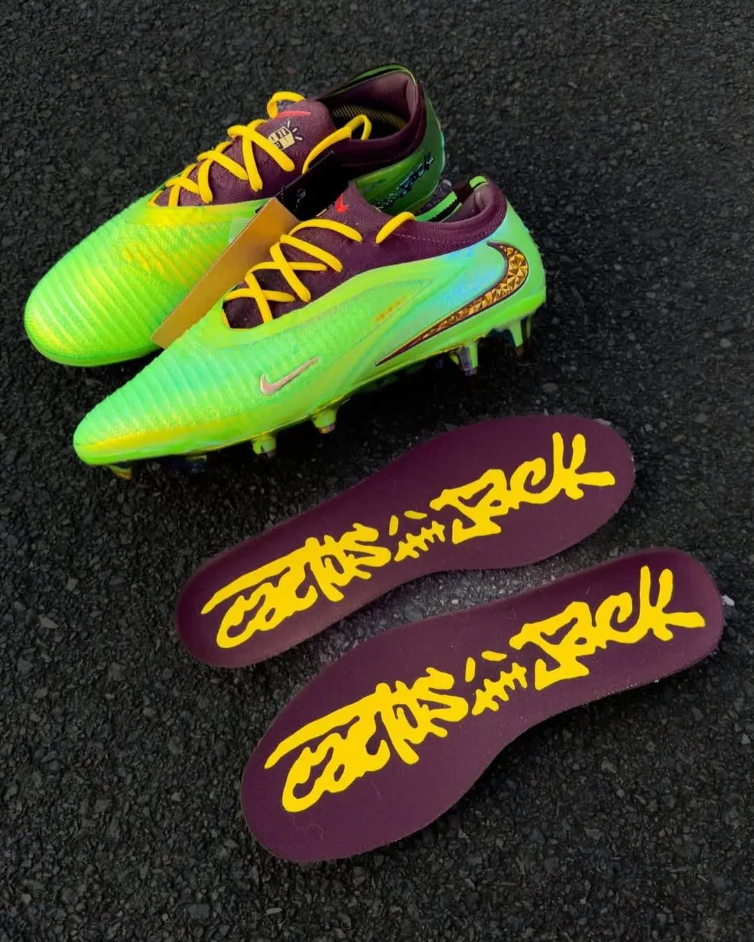 Product-style overhead image of a pair of bright football boots displayed on a dark textured surface alongside removable insoles. The boots feature a vivid neon green upper contrasted with deep burgundy interiors and bright yellow laces, while metallic detailing and textured side branding add a futuristic performance feel. The insoles are finished in burgundy with oversized handwritten-style Cactus Jack graphics in yellow, reinforcing the football-meets-streetwear collide aesthetic