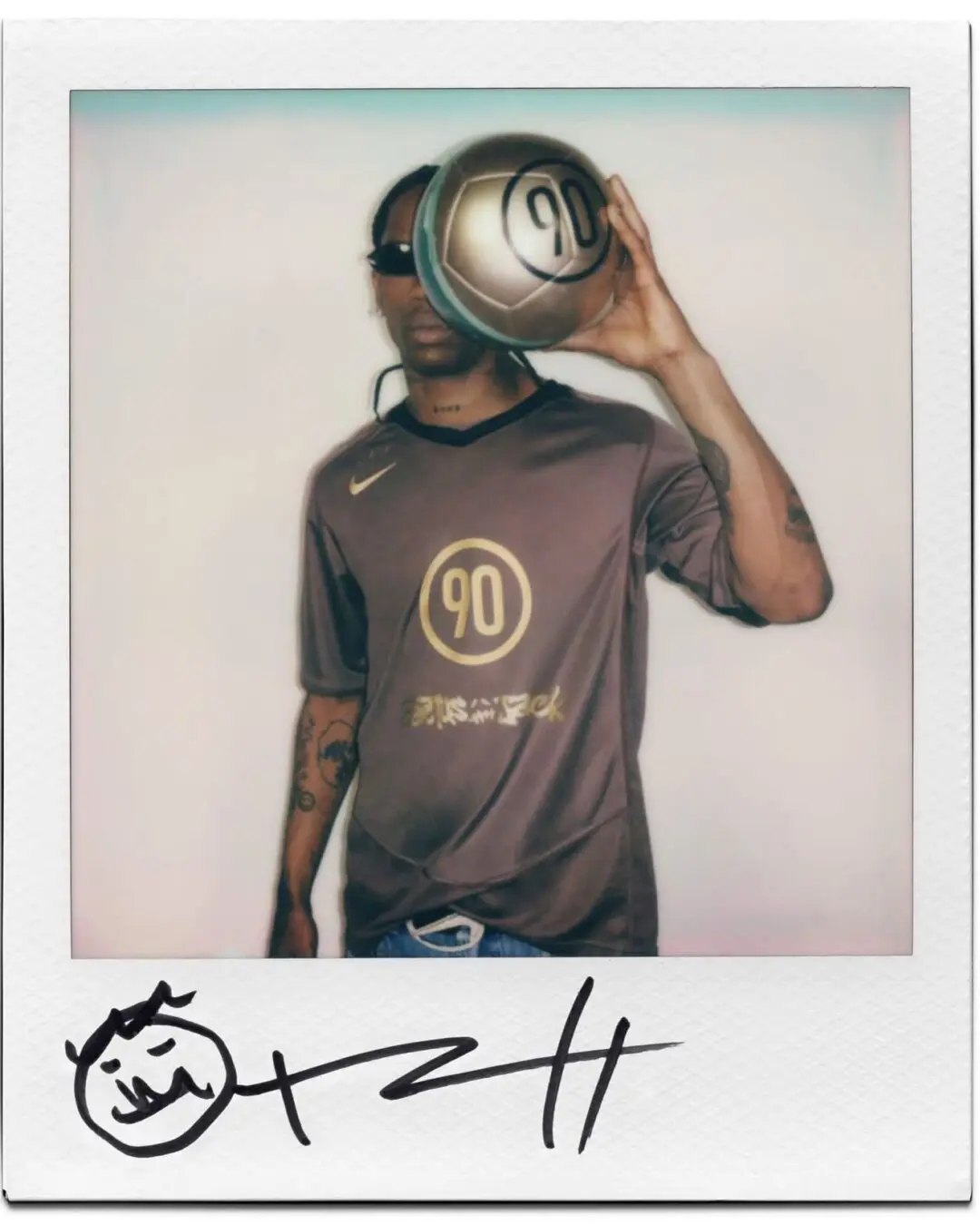 Polaroid-style campaign image showing a person in a dark football jersey with metallic gold graphics and a large “90” emblem centered on the chest while holding a matching gold soccer ball over one side of the face. The subject wears slim black sunglasses and stands against a minimal backdrop, while the instant-film border and hand-drawn doodle signature at the bottom create a nostalgic editorial and streetwear lookbook feel