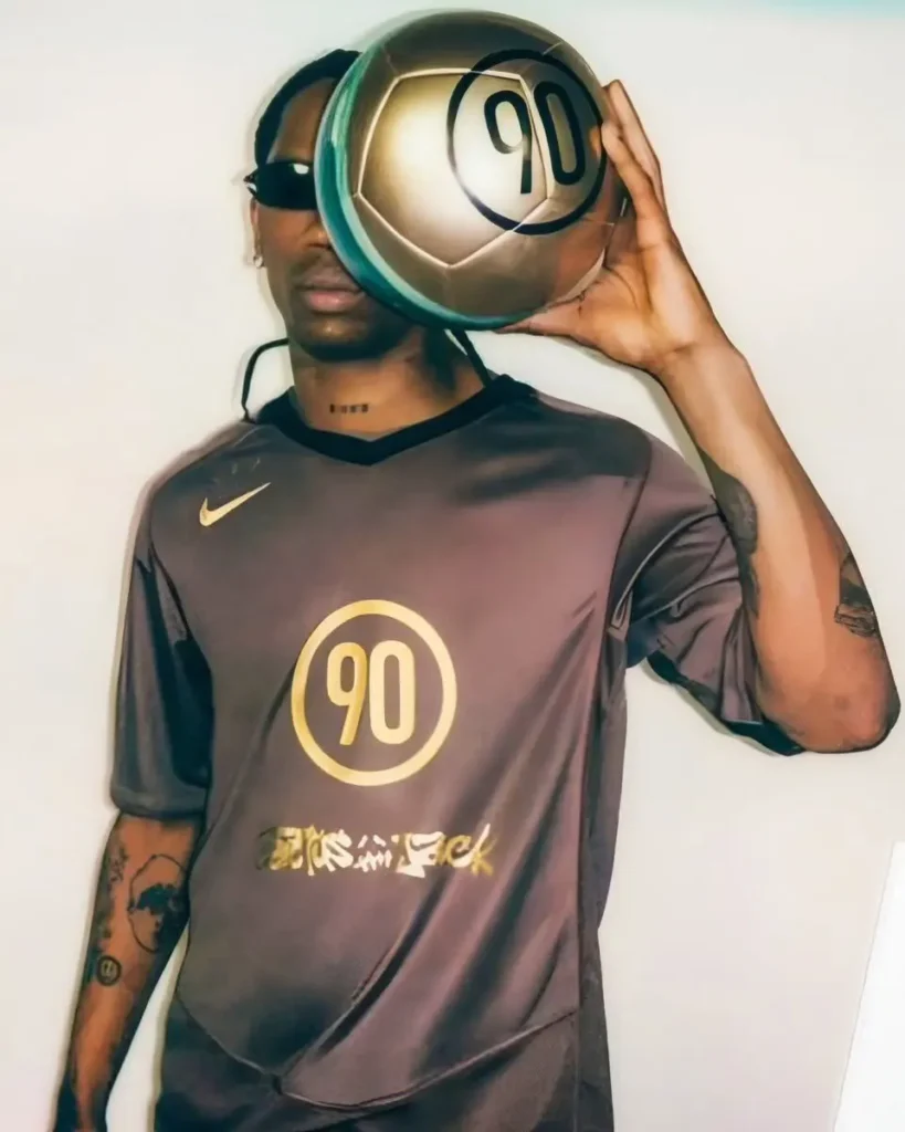 Editorial-style portrait of a person wearing a dark Nike football jersey with metallic gold detailing and a large “90” graphic at center while holding a matching gold soccer ball over one side of the face. The look is completed with narrow black sunglasses, visible tattoos, and minimalist studio lighting against a clean backdrop, giving the image a fashion campaign and football culture crossover feel