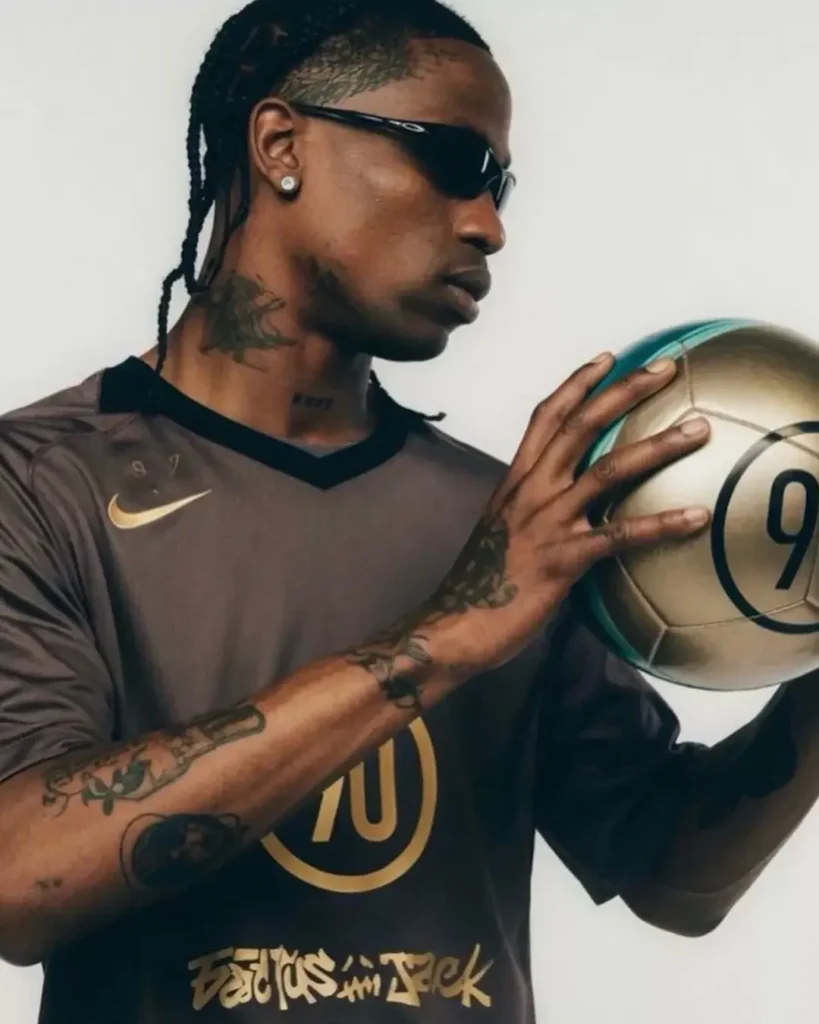 Close-up editorial portrait of a person in a dark Nike football jersey with metallic gold graphics, holding a matching gold soccer ball marked with a large “90.” Wearing narrow sport sunglasses and showcasing braided hair and visible tattoos, the subject is captured in profile against a minimal studio backdrop, emphasizing the collection’s fusion of football performance aesthetics and fashion campaign style