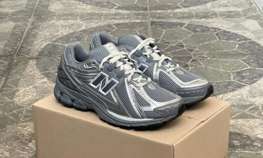 New Balance 1906R running shoe in a grey and silver colorway displayed atop a cardboard shoebox, featuring breathable mesh construction, metallic synthetic overlays, cream laces, and a sculpted N-ergy cushioned sole against a stone-tiled background